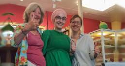 Gisela Zimmermann (Martha Café), Meryem und Inklusionsberaterin Stefanie Schreiner stehen (von links nach rechts) vor der Theke des Martha Cafés in Nürnberg und lächeln. Die drei Frauen strecken den Daumen nach oben als Zeichen für „gut gemacht“.